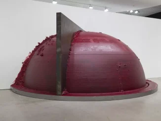 Grossformatige Skulptur: dunkelrote Halbkugel mit metallener Klinge, die die Oberfläche durchschneidet, Pigmentreste am Rand
