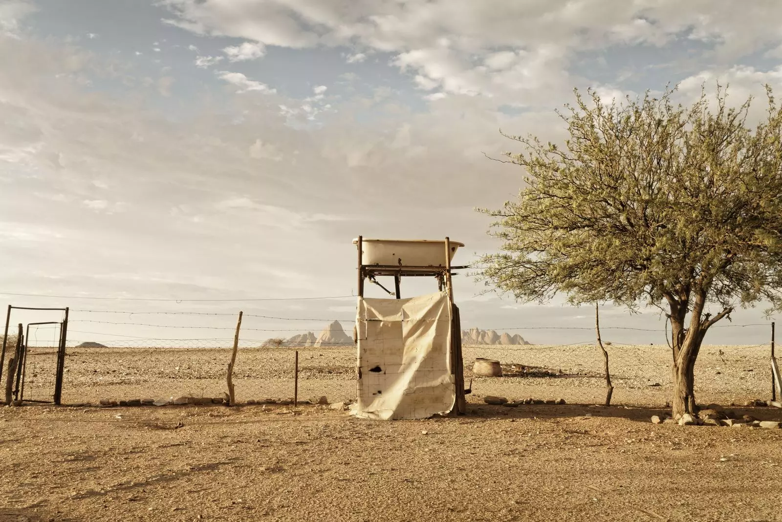 Geographies of Drought – Margaret Courtney-Clarke, Farbfotografie: karge Wüstenlandschaft mit verfallener Holzkonstruktion und einzelnem Baum