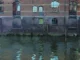 Lars Möller, Spiegelung, 2016: Gemaelde eines Backsteinlagerhauses am Wasser mit Spiegelung im dunklen Kanal
