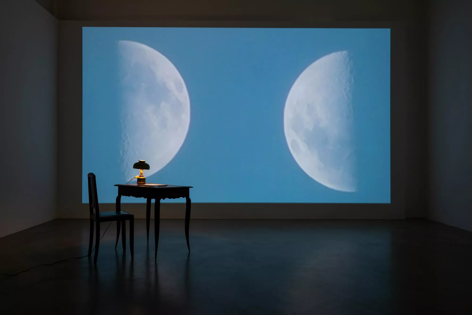Regina Hübner Ausstellung – Videoinstallation: Zwei Mondphasen auf blauer Projektionswand, davor antiker Tisch mit Öllampe und Stuhl