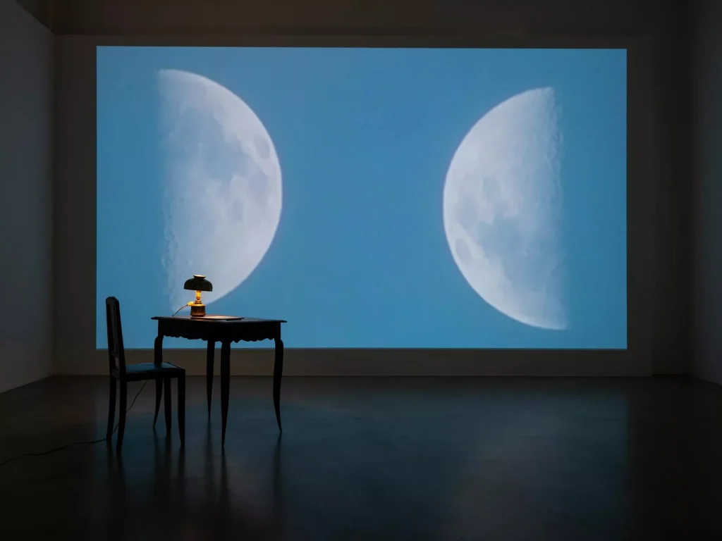 Videoinstallation: Zwei Mondphasen auf blauer Projektionswand, davor antiker Tisch mit Öllampe und Stuhl