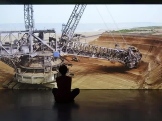 Silhouette einer sitzenden Person vor grossformatiger Videoprojektion eines Tagebau-Baggers in Erdlandschaft
