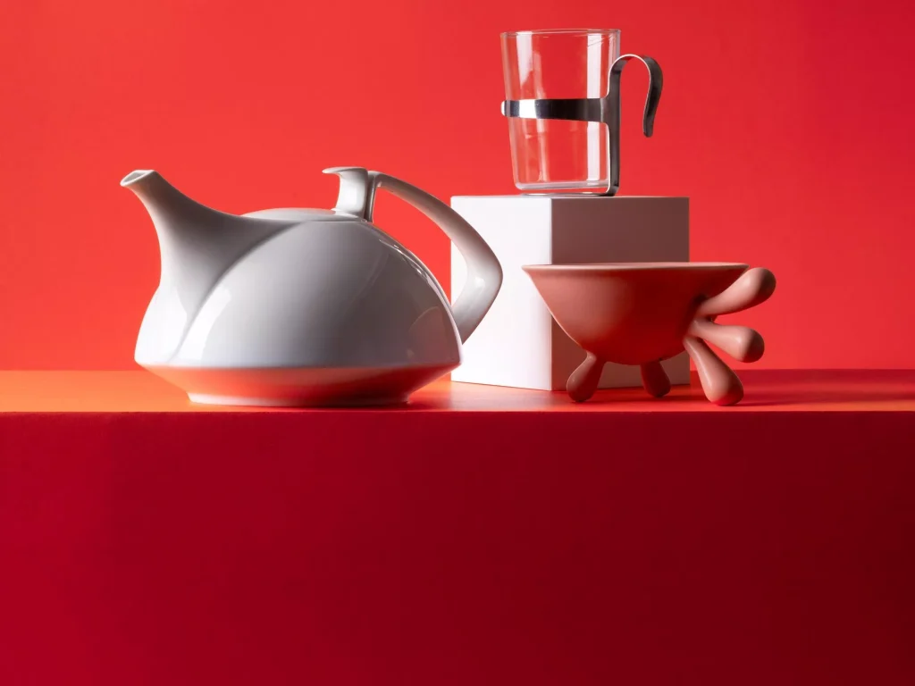 Designobjekte vor rotem Hintergrund: weisse Porzellan-Teekanne, Glasbecher mit Metallhalter und rote Schale auf organischen Fuesssen