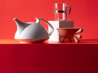 Designobjekte vor rotem Hintergrund: weisse Porzellan-Teekanne, Glasbecher mit Metallhalter und rote Schale auf organischen Fuesssen