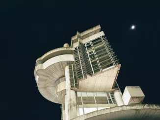 Tobias Zielony, Overshoot-2: Nachtaufnahme eines Brutalismus-Gebäudes von unten, Mond am schwarzen Himmel